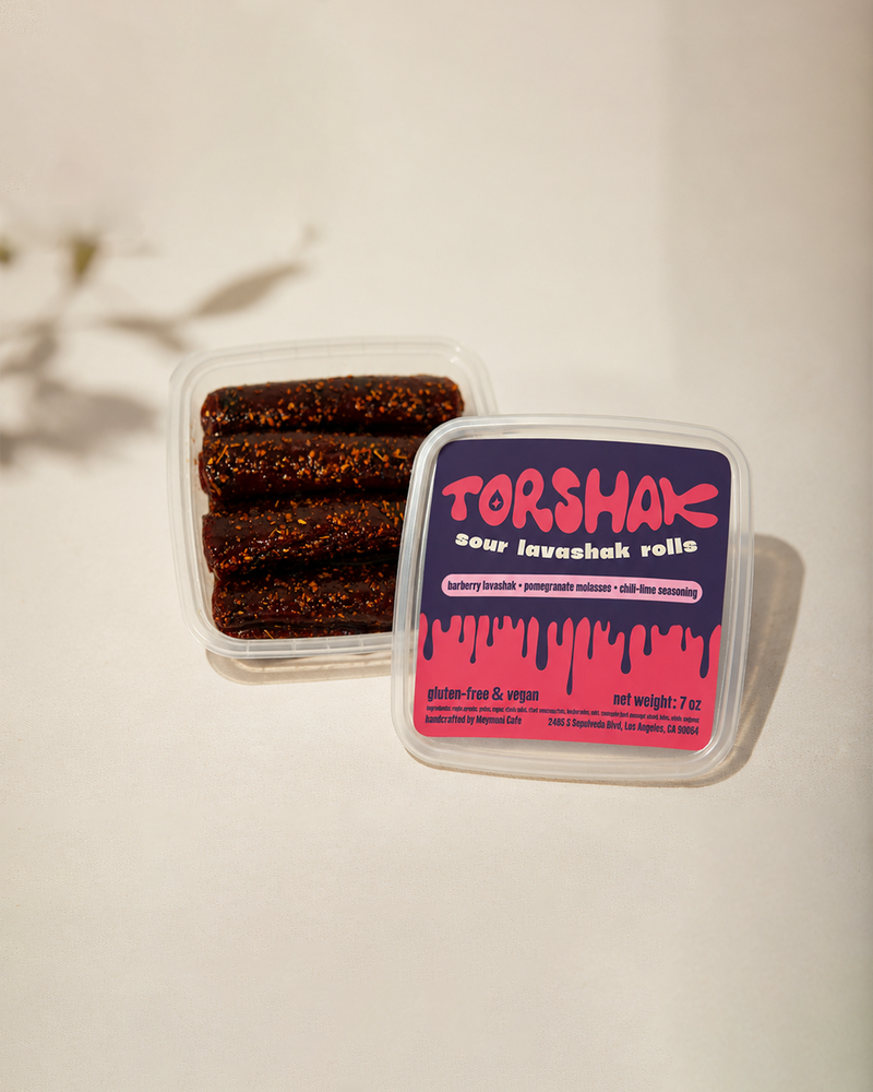 Two clear plastic tubs of Torshak sour lavashak rolls on a light beige background. One tub is open, showing three dark brown rolled pieces covered in coarse sesame seeds, and the other is closed with a dark purple and bright pink label featuring the Torshak brand name.